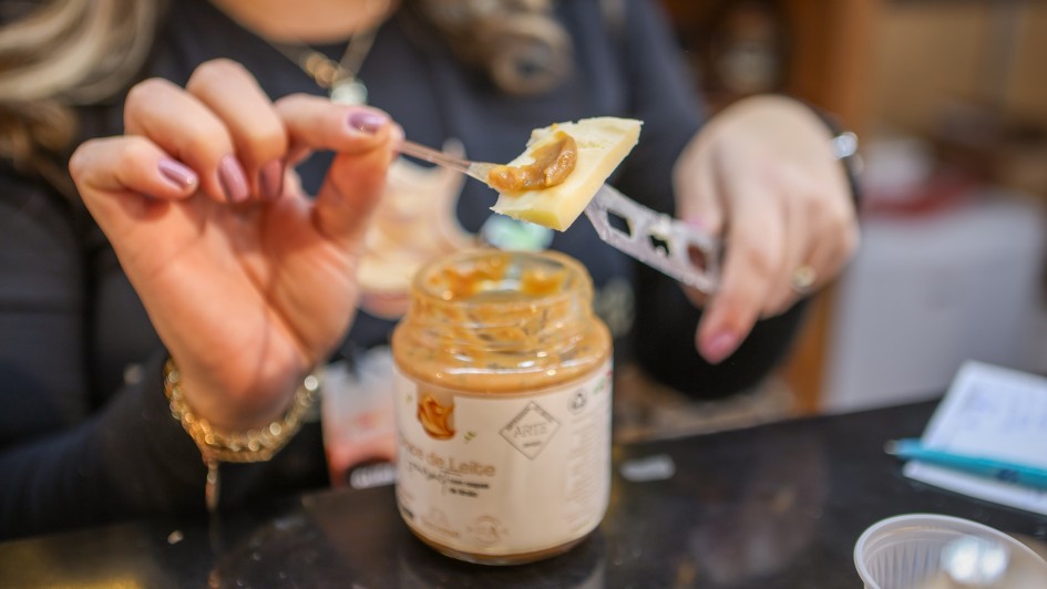Foto mostra o detalhe de um pote de doce de leite, em que uma m&atilde;o feminina est&aacute; servindo o doce sobre um peda&ccedil;o de queijo, que ela segura com uma faca. 