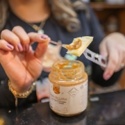 Foto mostra o detalhe de um pote de doce de leite, em que uma m&atilde;o feminina est&aacute; servindo o doce sobre um peda&ccedil;o de queijo, que ela segura com uma faca. 