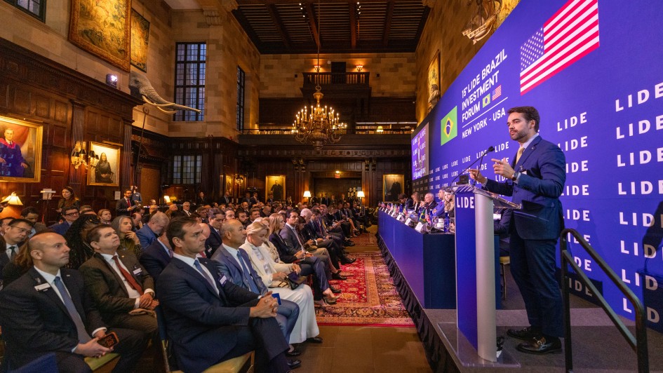 A fotografia, capturada em um plano geral e &acirc;ngulo lateral, mostra o governador Eduardo Leite discursando em um evento de investimentos em um sal&atilde;o imponente de arquitetura cl&aacute;ssica.

O Orador: &Agrave; direita da imagem, Eduardo Leite est&aacute; de p&eacute; atr&aacute;s de um p&uacute;lpito transparente com o logotipo "LIDE". Ele veste um terno azul-marinho e gesticula com as m&atilde;os enquanto fala ao microfone. Atr&aacute;s dele, h&aacute; um grande painel azul vibrante com as inscri&ccedil;&otilde;es "1&deg; LIDE BRAZIL INVESTMENT FORUM", as bandeiras do Brasil e dos EUA, e a localiza&ccedil;&atilde;o "NEW YORK - USA".

O P&uacute;blico: &Agrave; esquerda e ao centro, uma plateia numerosa composta majoritariamente por homens e mulheres em trajes formais (ternos e tailleurs) assiste ao discurso. Eles est&atilde;o sentados em cadeiras dispostas sobre tapetes orientais vermelhos que cobrem o ch&atilde;o de madeira.

A Mesa de Autoridades: Entre o orador e a plateia, h&aacute; uma mesa comprida coberta com uma toalha azul, onde diversas autoridades est&atilde;o sentadas de frente para o p&uacute;blico.

O Ambiente: O sal&atilde;o possui um p&eacute;-direito muito alto com paredes de pedra e detalhes em madeira escura. A decora&ccedil;&atilde;o &eacute; luxuosa, incluindo:

Grandes quadros de retratos a &oacute;leo com molduras douradas nas paredes.

Tape&ccedil;arias cl&aacute;ssicas penduradas na parte superior.

Um grande lustre de metal em estilo candelabro pendurado no centro do teto.

Uma curiosa cabe&ccedil;a de elefante esculpida (provavelmente parte da decora&ccedil;&atilde;o hist&oacute;rica do local) projetando-se da parede &agrave; esquerda.

Um mezanino com estantes de livros vis&iacute;vel ao fundo.

A imagem transmite a escala e a import&acirc;ncia de um f&oacute;rum internacional de neg&oacute;cios e pol&iacute;tica.