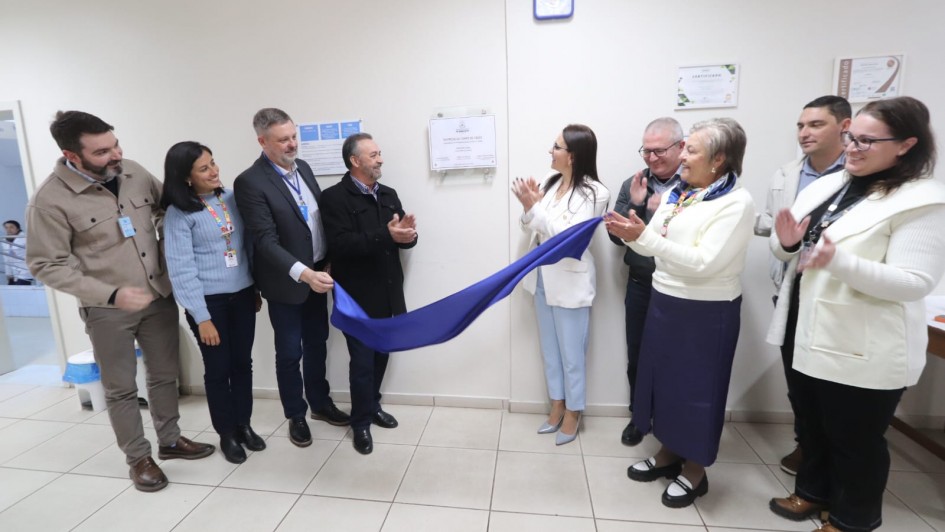 A fotografia em plano geral mostra um grupo de nove pessoas reunidas em um corredor hospitalar para a cerim&ocirc;nia de descerramento de uma placa inaugural.

A&ccedil;&atilde;o Central: No centro da imagem, um homem e uma mulher puxam simultaneamente um tecido de cetim azul, revelando uma placa comemorativa fixada na parede branca. As outras pessoas ao redor sorriem e aplaudem o momento.

Os Participantes:

&Agrave; esquerda, quatro pessoas observam: um homem com jaqueta bege, uma mulher com su&eacute;ter azul claro e crach&aacute; colorido, um homem de terno escuro auxiliando no descerramento e um homem de casaco preto tamb&eacute;m segurando o tecido.

&Agrave; direita, cinco pessoas acompanham: a mulher que ajuda a puxar o tecido veste um blazer branco e cal&ccedil;a azul clara; ao lado dela, um homem de &oacute;culos e uma mulher de blusa branca e saia longa azul marinho aplaudem; na extremidade direita, um homem de jaqueta clara e uma mulher de casaco branco completam o grupo.

Elementos da Parede:

A placa inaugural &eacute; retangular, de fundo branco, protegida por uma placa de acr&iacute;lico transparente presa por quatro parafusos met&aacute;licos.

Acima da placa, h&aacute; um rel&oacute;gio de parede quadrado com moldura azul.

&Agrave; direita, dois certificados emoldurados est&atilde;o pendurados na parede.

&Agrave; esquerda da placa, h&aacute; um pequeno cartaz azul com avisos ou instru&ccedil;&otilde;es.

Ambiente: O local possui paredes brancas, ilumina&ccedil;&atilde;o artificial clara e uniforme, e ch&atilde;o revestido com pisos cer&acirc;micos em tom bege. No canto inferior esquerdo, v&ecirc;-se um cesto de lixo branco com tampa azul.

A imagem registra um momento formal de celebra&ccedil;&atilde;o e entrega de melhorias institucionais.