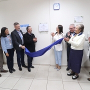 A fotografia em plano geral mostra um grupo de nove pessoas reunidas em um corredor hospitalar para a cerim&ocirc;nia de descerramento de uma placa inaugural.

A&ccedil;&atilde;o Central: No centro da imagem, um homem e uma mulher puxam simultaneamente um tecido de cetim azul, revelando uma placa comemorativa fixada na parede branca. As outras pessoas ao redor sorriem e aplaudem o momento.

Os Participantes:

&Agrave; esquerda, quatro pessoas observam: um homem com jaqueta bege, uma mulher com su&eacute;ter azul claro e crach&aacute; colorido, um homem de terno escuro auxiliando no descerramento e um homem de casaco preto tamb&eacute;m segurando o tecido.

&Agrave; direita, cinco pessoas acompanham: a mulher que ajuda a puxar o tecido veste um blazer branco e cal&ccedil;a azul clara; ao lado dela, um homem de &oacute;culos e uma mulher de blusa branca e saia longa azul marinho aplaudem; na extremidade direita, um homem de jaqueta clara e uma mulher de casaco branco completam o grupo.

Elementos da Parede:

A placa inaugural &eacute; retangular, de fundo branco, protegida por uma placa de acr&iacute;lico transparente presa por quatro parafusos met&aacute;licos.

Acima da placa, h&aacute; um rel&oacute;gio de parede quadrado com moldura azul.

&Agrave; direita, dois certificados emoldurados est&atilde;o pendurados na parede.

&Agrave; esquerda da placa, h&aacute; um pequeno cartaz azul com avisos ou instru&ccedil;&otilde;es.

Ambiente: O local possui paredes brancas, ilumina&ccedil;&atilde;o artificial clara e uniforme, e ch&atilde;o revestido com pisos cer&acirc;micos em tom bege. No canto inferior esquerdo, v&ecirc;-se um cesto de lixo branco com tampa azul.

A imagem registra um momento formal de celebra&ccedil;&atilde;o e entrega de melhorias institucionais.
