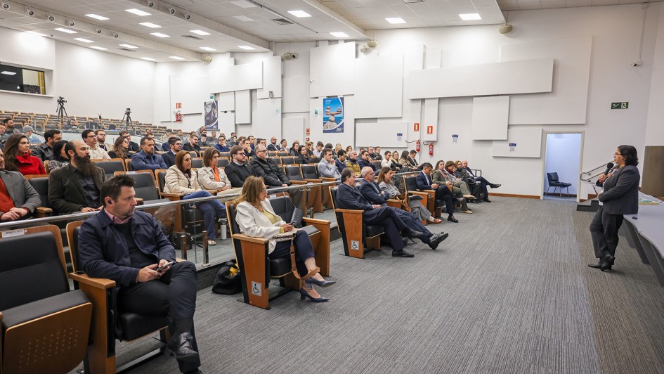 Esta &eacute; uma descri&ccedil;&atilde;o da imagem "Governo do Estado promove semin&aacute;rio com debate sobre os impactos fiscais da reforma tribut&aacute;ria.jpg" para fins de acessibilidade:

A fotografia, capturada em um plano geral e &acirc;ngulo lateral, mostra um audit&oacute;rio moderno durante a realiza&ccedil;&atilde;o de um semin&aacute;rio.

A Palestrante: &Agrave; direita da imagem, uma mulher com cabelos escuros e &oacute;culos, vestindo um conjunto formal de blazer e cal&ccedil;as cinza-escuro, fala ao microfone. Ela est&aacute; de p&eacute;, voltada para a plateia, posicionada &agrave; frente de uma mesa comprida de cor azulada que comp&otilde;e o palco.

O P&uacute;blico: O audit&oacute;rio est&aacute; ocupado por diversas pessoas sentadas em poltronas pretas com acabamento em madeira. Os participantes, homens e mulheres de v&aacute;rias idades, vestem trajes sociais e casuais, muitos deles voltados com aten&ccedil;&atilde;o para a palestrante. Algumas poltronas de ponta de fileira possuem o s&iacute;mbolo de acessibilidade para cadeirantes.

O Ambiente:

As poltronas est&atilde;o organizadas em fileiras em um plano inclinado (estilo anfiteatro).

As paredes s&atilde;o brancas e decoradas com pain&eacute;is retangulares em relevo, de diferentes tamanhos, que conferem uma textura moderna e auxiliam na ac&uacute;stica.

O teto &eacute; composto por placas modulares brancas com ilumina&ccedil;&atilde;o embutida por lumin&aacute;rias quadradas e spots direcionais.

O ch&atilde;o &eacute; revestido por um carpete cinza com listras sutis.

Ao fundo, &agrave; esquerda, &eacute; poss&iacute;vel ver um trip&eacute; com uma c&acirc;mera fotogr&aacute;fica ou de v&iacute;deo montada para registrar o evento.

&Agrave; direita, h&aacute; uma porta de sa&iacute;da aberta que revela um pequeno espa&ccedil;o interno com uma cadeira.

A imagem transmite um ambiente de di&aacute;logo t&eacute;cnico, profissionalismo e troca de conhecimentos em um evento institucional.