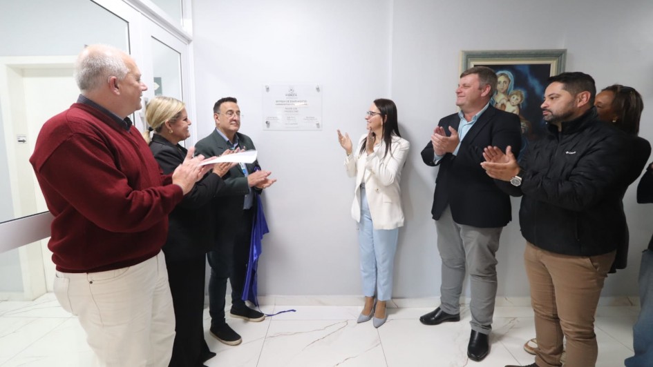 A imagem mostra um ato de inaugura&ccedil;&atilde;o em ambiente interno, com grupo reunido diante de uma placa de identifica&ccedil;&atilde;o fixada na parede, sendo revelada com faixa. As pessoas est&atilde;o dispostas ao redor e aplaudem ap&oacute;s a cerim&ocirc;nia.