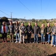 Registro coletivo em horta comunit&aacute;ria, com grupo alinhado em &aacute;rea de cultivo ao ar livre. Ao fundo, h&aacute; canteiros com plantas, casas e rede el&eacute;trica.