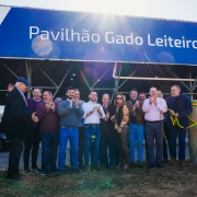 Registro coletivo em frente ao Pavilh&atilde;o Gado Leiteiro, durante ato oficial com corte simb&oacute;lico de fita nas laterais. Um grupo est&aacute; alinhado na entrada, com estrutura met&aacute;lica do galp&atilde;o ao fundo e luz do sol incidindo sobre a cena, marcando a abertura do espa&ccedil;o expositivo.