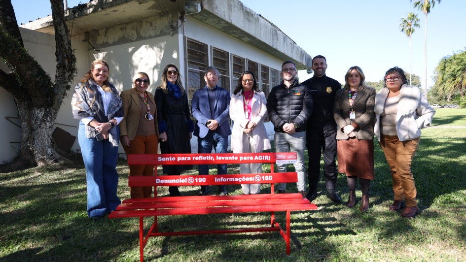 Esta &eacute; uma descri&ccedil;&atilde;o da imagem "Arthur Pl&aacute;cido Ascom SSPS (3).jpg" para fins de acessibilidade:

A fotografia mostra um grupo de nove pessoas posando em p&eacute; atr&aacute;s de um banco de pra&ccedil;a vermelho, em uma &aacute;rea externa gramada e ensolarada.

O Banco Vermelho: Posicionado em primeiro plano, o banco met&aacute;lico de cor vermelha vibrante &eacute; o elemento central. Ele possui frases de conscientiza&ccedil;&atilde;o pintadas em branco em seus tr&ecirc;s sarrafos do encosto:

No sarrafo superior: "Sente para refletir, levante para AGIR!".

No sarrafo central: "Denuncie! (&iacute;cone de telefone) 190".

No sarrafo inferior: "Informa&ccedil;&otilde;es: (&iacute;cone de telefone) 180".

O Grupo de Pessoas: Nove adultos (seis mulheres e tr&ecirc;s homens) est&atilde;o alinhados lado a lado atr&aacute;s do banco. Eles vestem roupas casuais e formais de inverno, como jaquetas, blazers, sobretudos e cachec&oacute;is. Muitos utilizam crach&aacute;s com cord&otilde;es coloridos (verde, vermelho e amarelo). Entre eles, destaca-se um homem ao centro-direita vestindo um uniforme operacional preto com um bras&atilde;o no peito e outro homem ao lado vestindo uma jaqueta acolchoada com a bandeira do Rio Grande do Sul.

O Ambiente:

O grupo est&aacute; sobre um gramado verde, com sombras n&iacute;tidas projetadas pelo sol forte.

Ao fundo, &agrave; esquerda, h&aacute; uma &aacute;rvore de tronco grosso e uma edifica&ccedil;&atilde;o simples de paredes brancas e janelas com persianas externas de madeira.

&Agrave; direita, o gramado se estende sob um c&eacute;u azul limpo, com palmeiras altas vis&iacute;veis ao longe.

A imagem registra uma a&ccedil;&atilde;o institucional de conscientiza&ccedil;&atilde;o e combate &agrave; viol&ecirc;ncia, possivelmente relacionada a campanhas de prote&ccedil;&atilde;o &agrave; mulher.