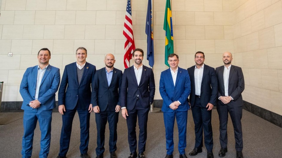 A fotografia mostra sete homens posando em p&eacute;, lado a lado, em um ambiente interno amplo com est&eacute;tica institucional.

O Grupo:

No centro da imagem est&aacute; o governador Eduardo Leite, um homem branco de barba e cabelos castanhos curtos, vestindo um terno azul-marinho sobre camisa branca sem gravata.

Ele est&aacute; ladeado por outros seis homens, todos brancos e vestindo trajes sociais (ternos em tons de azul e cinza-escuro com camisas claras).

Todos os integrantes do grupo sorriem para a c&acirc;mera e mant&ecirc;m uma postura formal, a maioria com as m&atilde;os cruzadas &agrave; frente do corpo.

Plano de Fundo:

Logo atr&aacute;s de Eduardo Leite, encontram-se tr&ecirc;s bandeiras em seus respectivos mastros: a bandeira dos Estados Unidos (&agrave; esquerda), uma bandeira azul (ao centro) e a bandeira do Brasil (&agrave; direita).

A parede ao fundo &eacute; composta por grandes blocos de pedra clara ou cer&acirc;mica, conferindo um aspecto s&oacute;brio e moderno ao local.

O Ambiente:

O ch&atilde;o &eacute; revestido por um carpete cinza escuro uniforme.

A ilumina&ccedil;&atilde;o &eacute; clara e distribu&iacute;da por igual em toda a cena.

A imagem registra um encontro oficial de natureza diplom&aacute;tica ou de neg&oacute;cios entre representantes governamentais e investidores.