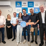 A fotografia mostra um grupo de sete pessoas sorridentes em um ato oficial de entrega de chaves, em um ambiente interno.

A&ccedil;&atilde;o Central: No centro, o governador Eduardo Leite e um homem &agrave; sua direita seguram juntos uma chave simb&oacute;lica gigante feita de material r&iacute;gido. A chave &eacute; colorida (azul, laranja e branco) e cont&eacute;m o texto "ENTREGA DO VILLA PIENZA RESIDENCIAL". Entre eles, uma mulher jovem, de cabelos cacheados, segura um molho de chaves real com um chaveiro vermelho, representando a benefici&aacute;ria do programa.

Os Participantes:

Eduardo Leite (ao centro) veste uma camisa polo azul-marinho, cal&ccedil;a jeans escura e t&ecirc;nis claros.

&Agrave; esquerda da benefici&aacute;ria, tr&ecirc;s pessoas observam: um homem de camisa social azul clara, uma mulher de blusa preta e um homem de camisa rosa clara.

&Agrave; direita do grupo central, al&eacute;m do homem que segura a chave gigante (de camisa preta e bra&ccedil;o tatuado), h&aacute; um homem de terno preto e cabelos grisalhos que sorri abertamente para a c&acirc;mera.

Plano de Fundo: Atr&aacute;s do grupo, h&aacute; um painel (back-drop) com um padr&atilde;o de quadrados azuis e brancos contendo logotipos oficiais: "Minha Casa Minha Vida", "Porta de Entrada", "Caixa FGTS" e o logotipo do Governo Federal (Brasil).

O Ambiente: A sala tem paredes brancas e um aparelho de ar-condicionado do tipo split instalado no alto, &agrave; esquerda. O ch&atilde;o &eacute; de piso cer&acirc;mico claro.

A imagem registra um momento de celebra&ccedil;&atilde;o pela entrega de moradias populares e parcerias habitacionais.