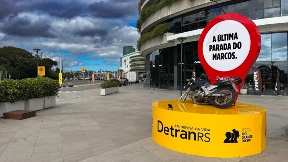 A imagem mostra uma instala&ccedil;&atilde;o urbana em &aacute;rea de circula&ccedil;&atilde;o p&uacute;blica, com uma motocicleta posicionada sobre uma base circular amarela com o texto &ldquo;DetranRS em defesa da vida&rdquo; e marca do Governo do Estado do Rio Grande do Sul. Atr&aacute;s da moto, h&aacute; um grande elemento visual em formato de marcador com fundo branco e borda vermelha, contendo a frase &ldquo;A &uacute;ltima parada do Marcos.&rdquo;