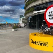 A imagem mostra uma instala&ccedil;&atilde;o urbana em &aacute;rea de circula&ccedil;&atilde;o p&uacute;blica, com uma motocicleta posicionada sobre uma base circular amarela com o texto &ldquo;DetranRS em defesa da vida&rdquo; e marca do Governo do Estado do Rio Grande do Sul. Atr&aacute;s da moto, h&aacute; um grande elemento visual em formato de marcador com fundo branco e borda vermelha, contendo a frase &ldquo;A &uacute;ltima parada do Marcos.&rdquo;