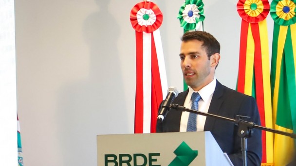 A fotografia em plano m&eacute;dio mostra um homem discursando em um ambiente institucional formal.

O Orador: Um homem branco, jovem, de cabelos curtos escuros e barba por fazer bem aparada. Ele veste um terno azul-marinho, camisa social branca e uma gravata azul. Ele est&aacute; posicionado atr&aacute;s de um p&uacute;lpito, falando ao microfone com uma express&atilde;o s&eacute;ria e focada.

O P&uacute;lpito: O p&uacute;lpito &eacute; cinza-claro e exibe na parte frontal o logotipo do BRDE em letras pretas. Logo abaixo, l&ecirc;-se a inscri&ccedil;&atilde;o: "BANCO REGIONAL DE DESENVOLVIMENTO DO EXTREMO SUL". Ao lado da sigla, h&aacute; um s&iacute;mbolo geom&eacute;trico em tons de verde que remete ao mapa da regi&atilde;o Sul do Brasil.

Plano de Fundo: Atr&aacute;s do orador, veem-se os mastros de quatro bandeiras. No topo de cada mastro, h&aacute; rosetas e faixas decorativas coloridas que representam os estados de atua&ccedil;&atilde;o do banco:

Vermelho e branco (Paran&aacute;).

Verde e branco (Santa Catarina).

Duas faixas em verde, vermelho e amarelo (Rio Grande do Sul).

Ambiente: A parede ao fundo &eacute; lisa e de cor cinza-clara, com ilumina&ccedil;&atilde;o uniforme que destaca o orador e a marca da institui&ccedil;&atilde;o.

A imagem registra um momento de pronunciamento oficial, transmitindo credibilidade e profissionalismo.