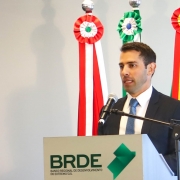 A fotografia em plano m&eacute;dio mostra um homem discursando em um ambiente institucional formal.

O Orador: Um homem branco, jovem, de cabelos curtos escuros e barba por fazer bem aparada. Ele veste um terno azul-marinho, camisa social branca e uma gravata azul. Ele est&aacute; posicionado atr&aacute;s de um p&uacute;lpito, falando ao microfone com uma express&atilde;o s&eacute;ria e focada.

O P&uacute;lpito: O p&uacute;lpito &eacute; cinza-claro e exibe na parte frontal o logotipo do BRDE em letras pretas. Logo abaixo, l&ecirc;-se a inscri&ccedil;&atilde;o: "BANCO REGIONAL DE DESENVOLVIMENTO DO EXTREMO SUL". Ao lado da sigla, h&aacute; um s&iacute;mbolo geom&eacute;trico em tons de verde que remete ao mapa da regi&atilde;o Sul do Brasil.

Plano de Fundo: Atr&aacute;s do orador, veem-se os mastros de quatro bandeiras. No topo de cada mastro, h&aacute; rosetas e faixas decorativas coloridas que representam os estados de atua&ccedil;&atilde;o do banco:

Vermelho e branco (Paran&aacute;).

Verde e branco (Santa Catarina).

Duas faixas em verde, vermelho e amarelo (Rio Grande do Sul).

Ambiente: A parede ao fundo &eacute; lisa e de cor cinza-clara, com ilumina&ccedil;&atilde;o uniforme que destaca o orador e a marca da institui&ccedil;&atilde;o.

A imagem registra um momento de pronunciamento oficial, transmitindo credibilidade e profissionalismo.