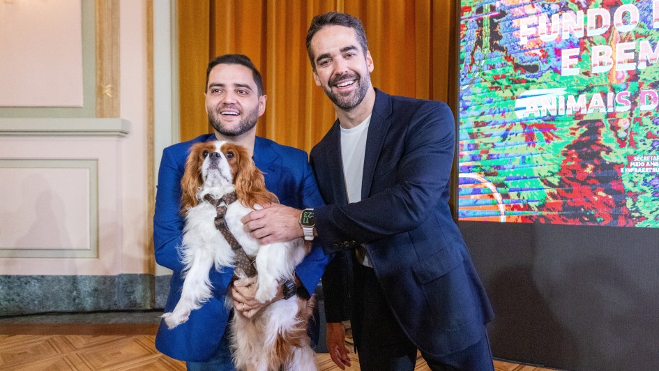Registro institucional em ambiente interno, com duas pessoas posicionadas lado a lado enquanto uma delas segura um cachorro de pequeno porte com pelagem branca e marrom. Ao fundo, h&aacute; um painel digital colorido com elementos gr&aacute;ficos e texto relacionado a fundo de prote&ccedil;&atilde;o e bem-estar animal.