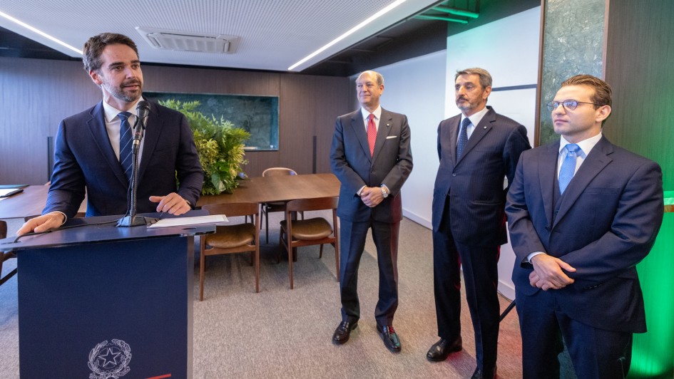 Discurso em ambiente institucional, com o governador posicionado atr&aacute;s de um p&uacute;lpito com emblema oficial e elemento gr&aacute;fico com cores da It&aacute;lia. Ao lado, outras pessoas permanecem em p&eacute; acompanhando a fala. 