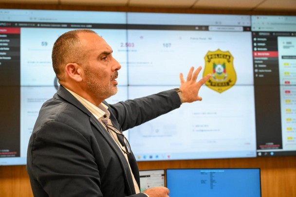 Em um ambiente tecnológico de monitoramento policial, o  cel. Alex Sandre Pinheiro Severo, diretor do DCCI, um homem de terno gesticula diante de um grande painel digital repleto de gráficos, indicadores. 