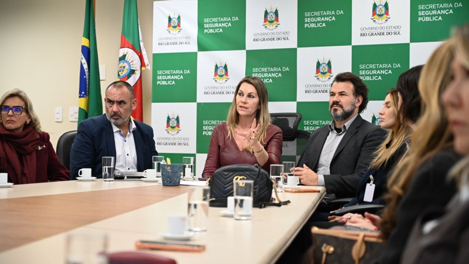 Em uma reuni&atilde;o oficial, v&aacute;rios participantes est&atilde;o sentados em torno de uma mesa de confer&ecirc;ncia, com copos, cadernos e objetos pessoais sobre a superf&iacute;cie. Ao fundo, um painel institucional exibe o nome da Secretaria da Seguran&ccedil;a P&uacute;blica e o bras&atilde;o do Estado, acompanhado das bandeiras do Brasil e do Rio Grande do Sul. A cena reflete um encontro administrativo voltado para pol&iacute;ticas de seguran&ccedil;a p&uacute;blica.