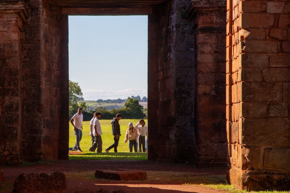 Vista através de uma estrutura histórica de pedra, formando um enquadramento natural para a paisagem externa. Ao fundo, um grupo caminha por área gramada sob luz natural, com árvores e relevo rural visíveis ao horizonte.