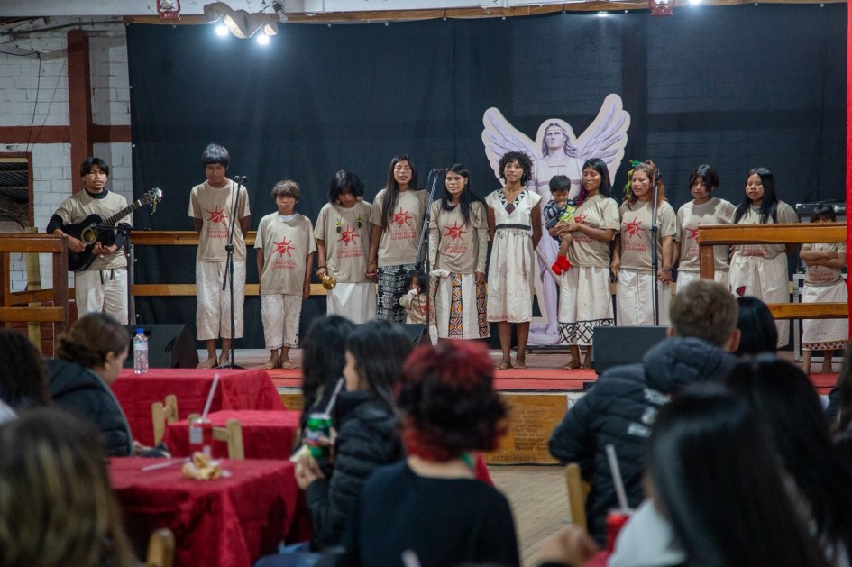 Apresentação cultural em espaço interno, com grupo alinhado em palco utilizando camisetas com a inscrição “Consciência Guarani”. Ao fundo, há cenário teatral com figura de anjo e microfones, enquanto o público acompanha sentado em mesas.