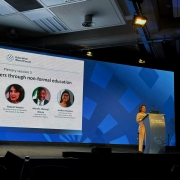 Painel em palco do Education World Forum, com tel&atilde;o exibindo a sess&atilde;o &ldquo;Plenary session 5 &ndash; Empowering learners through non-formal education&rdquo;. O slide apresenta fotos e nomes de participantes internacionais. &Agrave; direita do palco, a secret&aacute;ria da Educa&ccedil;&atilde;o est&aacute; em p&uacute;lpito com microfones e laptop, enquanto outro participante est&aacute; sentado &agrave; esquerda. 