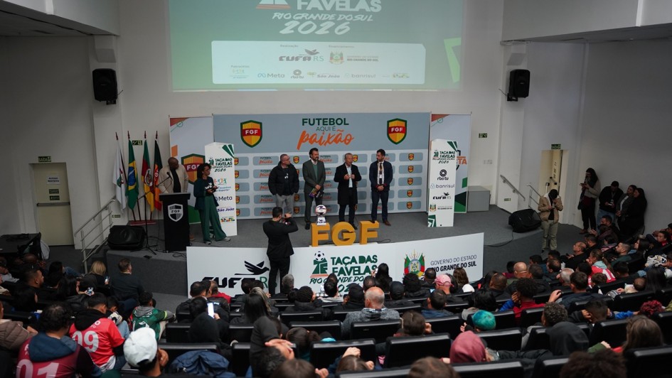 A fotografia, capturada em um plano geral e &acirc;ngulo elevado, mostra um audit&oacute;rio lotado durante o evento de lan&ccedil;amento da Ta&ccedil;a das Favelas Rio Grande do Sul 2026.

O Palco: No centro do palco elevado, quatro homens vestindo trajes sociais e casuais est&atilde;o alinhados lado a lado. Um deles, ao centro-direita, fala ao microfone.

&Agrave; esquerda do grupo principal, uma mulher negra veste um terno verde e segura pap&eacute;is, atuando como mestre de cerim&ocirc;nias ao lado de um p&uacute;lpito preto com o escudo da FGF (Federa&ccedil;&atilde;o Ga&uacute;cha de Futebol). No ch&atilde;o, &agrave; frente deles, destacam-se grandes letras tridimensionais amarelas com a sigla "FGF" e uma bola de futebol sobre um pedestal.

Um fot&oacute;grafo, de costas para a plateia, est&aacute; posicionado logo abaixo do palco registrando as autoridades.

Elementos Visuais e Identifica&ccedil;&atilde;o:

Ao fundo do palco, um grande painel exibe a frase "FUTEBOL AQUI &Eacute; paix&atilde;o" ladeada por escudos da FGF.

Acima do palco, uma grande tela de proje&ccedil;&atilde;o exibe a identidade oficial do evento com o t&iacute;tulo "TA&Ccedil;A DAS FAVELAS RIO GRANDE DO SUL 2026", destacando os logotipos da realiza&ccedil;&atilde;o pela CUFA RS (Central &Uacute;nica das Favelas) e financiamento do Governo do Estado do Rio Grande do Sul, al&eacute;m de patrocinadores como Meta, RBS TV e Banrisul.

Na base do palco, pain&eacute;is brancos repetem as marcas da CUFA, da Ta&ccedil;a das Favelas e do Governo do Estado.

No canto esquerdo do palco, est&atilde;o posicionadas quatro bandeiras oficiais em seus mastros.

O P&uacute;blico: O audit&oacute;rio em estilo anfiteatro est&aacute; repleto de pessoas sentadas em poltronas pretas, vistas de costas a partir do primeiro plano. Diversos jovens vestem camisetas esportivas coloridas (como uma vermelha com o n&uacute;mero 19 e outra com o n&uacute;mero 01), sugerindo a presen&ccedil;a de atletas ou representantes de comunidades.

A imagem transmite a atmosfera vibrante, institucional e comunit&aacute;ria de um grande evento de impacto social atrav&eacute;s do esporte.