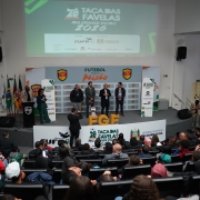 A fotografia, capturada em um plano geral e &acirc;ngulo elevado, mostra um audit&oacute;rio lotado durante o evento de lan&ccedil;amento da Ta&ccedil;a das Favelas Rio Grande do Sul 2026.

O Palco: No centro do palco elevado, quatro homens vestindo trajes sociais e casuais est&atilde;o alinhados lado a lado. Um deles, ao centro-direita, fala ao microfone.

&Agrave; esquerda do grupo principal, uma mulher negra veste um terno verde e segura pap&eacute;is, atuando como mestre de cerim&ocirc;nias ao lado de um p&uacute;lpito preto com o escudo da FGF (Federa&ccedil;&atilde;o Ga&uacute;cha de Futebol). No ch&atilde;o, &agrave; frente deles, destacam-se grandes letras tridimensionais amarelas com a sigla "FGF" e uma bola de futebol sobre um pedestal.

Um fot&oacute;grafo, de costas para a plateia, est&aacute; posicionado logo abaixo do palco registrando as autoridades.

Elementos Visuais e Identifica&ccedil;&atilde;o:

Ao fundo do palco, um grande painel exibe a frase "FUTEBOL AQUI &Eacute; paix&atilde;o" ladeada por escudos da FGF.

Acima do palco, uma grande tela de proje&ccedil;&atilde;o exibe a identidade oficial do evento com o t&iacute;tulo "TA&Ccedil;A DAS FAVELAS RIO GRANDE DO SUL 2026", destacando os logotipos da realiza&ccedil;&atilde;o pela CUFA RS (Central &Uacute;nica das Favelas) e financiamento do Governo do Estado do Rio Grande do Sul, al&eacute;m de patrocinadores como Meta, RBS TV e Banrisul.

Na base do palco, pain&eacute;is brancos repetem as marcas da CUFA, da Ta&ccedil;a das Favelas e do Governo do Estado.

No canto esquerdo do palco, est&atilde;o posicionadas quatro bandeiras oficiais em seus mastros.

O P&uacute;blico: O audit&oacute;rio em estilo anfiteatro est&aacute; repleto de pessoas sentadas em poltronas pretas, vistas de costas a partir do primeiro plano. Diversos jovens vestem camisetas esportivas coloridas (como uma vermelha com o n&uacute;mero 19 e outra com o n&uacute;mero 01), sugerindo a presen&ccedil;a de atletas ou representantes de comunidades.

A imagem transmite a atmosfera vibrante, institucional e comunit&aacute;ria de um grande evento de impacto social atrav&eacute;s do esporte.