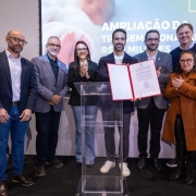 A fotografia em plano m&eacute;dio mostra um grupo de cinco pessoas (tr&ecirc;s homens e duas mulheres) posando em p&eacute;, lado a lado, em um ambiente interno iluminado.

Os Integrantes: Todos est&atilde;o sorrindo e voltados de frente para a c&acirc;mera.

Da esquerda para a direita:

O primeiro homem veste um palet&oacute; azul-marinho sobre camisa social branca e cal&ccedil;a jeans escura.

A primeira mulher tem cabelos loiros curtos, veste um casaco bege texturizado e cal&ccedil;as pretas.

O homem ao centro &eacute; o governador Eduardo Leite, que veste um palet&oacute; azul-marinho sobre uma camiseta branca e cal&ccedil;a clara.

A segunda mulher usa &oacute;culos, tem cabelos escuros e veste um blazer azul-marinho sobre uma blusa branca.

O terceiro homem, na extremidade direita, usa &oacute;culos, tem cabelos grisalhos e veste um terno cinza-escuro com camisa clara.

Plano de Fundo: Atr&aacute;s do grupo, h&aacute; uma grande parede de vidro que revela um jardim externo com folhagens verdes e plantas ornamentais sob a luz do dia, criando um efeito de transpar&ecirc;ncia e luminosidade natural.

O Ambiente: O ch&atilde;o interno &eacute; revestido com placas grandes de piso cer&acirc;mico ou porcelanato cinza-claro polido, que reflete sutilmente a silhueta das pessoas e a luz do ambiente.

A imagem registra um encontro oficial ou parceria institucional entre representantes governamentais e de sa&uacute;de.