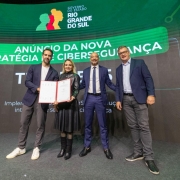 Esta &eacute; uma descri&ccedil;&atilde;o da imagem "Governo do Estado apresenta avan&ccedil;os do Programa RS+Digital e amplia parceria com munic&iacute;pios.jpeg" para fins de acessibilidade:

A fotografia em plano inteiro mostra quatro autoridades (tr&ecirc;s homens e uma mulher) alinhados lado a lado no centro de um palco, celebrando um an&uacute;ncio oficial.

A&ccedil;&atilde;o Central: &Agrave; esquerda, o governador Eduardo Leite e a mulher ao seu lado seguram juntos um documento oficial aberto em uma pasta de capa vermelha, exibindo-o para a c&acirc;mera. Todos os integrantes do grupo sorriem.

Os Participantes:

O governador Eduardo Leite (na extremidade esquerda) veste um palet&oacute; azul-marinho, camiseta branca, cal&ccedil;a cinza e t&ecirc;nis brancos.

A mulher ao seu lado veste um casaco longo de couro preto com saia plissada preta, meias escuras e botas pretas de cano m&eacute;dio.

O terceiro homem (centro-direita) veste um terno azul-vibrante com camisa branca, gravata escura e sapatos pretos sociais. Ele mant&eacute;m os bra&ccedil;os levemente abertos.

O quarto homem (na extremidade direita) usa &oacute;culos, veste um palet&oacute; azul-marinho sobre camisa azul-clara, cal&ccedil;a de sarja cinza e t&ecirc;nis escuros.

Plano de Fundo:

Atr&aacute;s do grupo, um grande tel&atilde;o de LED curvo exibe uma apresenta&ccedil;&atilde;o digital com fundo verde-escuro e detalhes gr&aacute;ficos.

No topo central do tel&atilde;o, destaca-se o logotipo oficial do Governo do Estado do Rio Grande do Sul (com o perfil de tr&ecirc;s rostos coloridos em verde, vermelho e amarelo).

Abaixo do logotipo, uma grande faixa verde-clara exibe em letras brancas o t&iacute;tulo: "AN&Uacute;NCIO DA NOVA ESTRAT&Eacute;GIA DE CIBERSEGURAN&Ccedil;A".

Mais abaixo,文字 brancas menores trazem textos informativos parcialmente vis&iacute;veis.

O Ambiente: O palco possui um revestimento cinza-escuro fosco e uniforme, com ilumina&ccedil;&atilde;o frontal direcionada para o grupo.

A imagem registra um momento institucional de avan&ccedil;o tecnol&oacute;gico e seguran&ccedil;a digital para o estado.