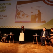 Esta &eacute; uma descri&ccedil;&atilde;o da imagem "Semin&aacute;rio do F&oacute;rum Permanente de Apoio &agrave; Forma&ccedil;&atilde;o de Professores fortalece pol&iacute;ticas de forma&ccedil;&atilde;o de professores no Estado.jpg" para fins de acessibilidade:

A fotografia em plano geral e &acirc;ngulo frontal mostra um painel de discuss&atilde;o em um palco de teatro ou audit&oacute;rio, com destaque para uma grande tela de proje&ccedil;&atilde;o ao fundo.

A Palestrante: No centro do palco, uma mulher de cabelos castanhos curtos est&aacute; de p&eacute; e fala ao microfone. Ela veste uma jaqueta de couro preta sobre blusa clara e uma saia longa plissada branca.

Os Integrantes do Painel:

&Agrave; direita, sentados em poltronas individuais de madeira com estofado escuro, tr&ecirc;s pessoas acompanham a apresenta&ccedil;&atilde;o: um homem de terno azul-escuro, camisa clara e gravata; uma mulher de casaco e saia escura com as pernas cruzadas; e uma mulher de jaqueta de couro preta e cal&ccedil;a escura.

&Agrave; esquerda, outras tr&ecirc;s pessoas tamb&eacute;m est&atilde;o acomodadas em poltronas semelhantes: um homem de palet&oacute; cinza que analisa pap&eacute;is em seu colo, uma mulher de blazer bord&ocirc; (parcialmente vis&iacute;vel na extremidade esquerda) e uma poltrona vazia com uma bolsa preta sobre o assento entre eles.

A Tela de Proje&ccedil;&atilde;o: O grande tel&atilde;o ao fundo exibe um slide institucional claro.

No lado esquerdo, l&ecirc;-se em letras mai&uacute;sculas douradas: "EDUCATION WORLD FORUM", seguido pelo nome "Sr. Olli-Pekka Heinonen" em marrom.

No centro, h&aacute; a reprodu&ccedil;&atilde;o de um slide em ingl&ecirc;s com o t&iacute;tulo "Education should function like a fractal", exibindo a silhueta de um homem discursando em um p&uacute;lpito e diagramas circulares interconectados.

No canto inferior direito do tel&atilde;o, consta o bras&atilde;o e o logotipo oficial do Governo do Estado do Rio Grande do Sul.

O Ambiente: O palco possui piso de t&aacute;buas de madeira claras e ilumina&ccedil;&atilde;o c&ecirc;nica direcionada. Pequenas mesas redondas de madeira servem de apoio para ta&ccedil;as e garrafas de &aacute;gua entre os participantes. O restante do cen&aacute;rio ao fundo &eacute; escuro.

A imagem registra um evento oficial e acad&ecirc;mico de relev&acirc;ncia internacional na &aacute;rea da educa&ccedil;&atilde;o.