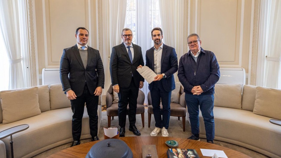 A imagem mostra quatro pessoas em p&eacute; em uma sala elegante, organizadas lado a lado em frente a uma grande janela com cortinas claras e luz natural entrando. Duas das pessoas ao centro seguram um documento, exibindo-o para a c&acirc;mera.