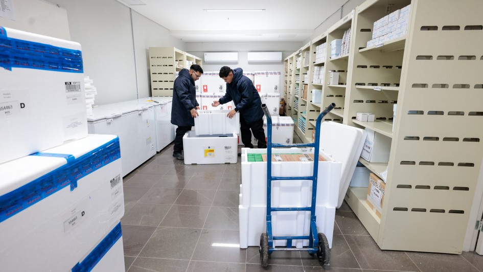 A imagem mostra um ambiente interno organizado, semelhante a uma sala de armazenamento refrigerado, com prateleiras met&aacute;licas claras nas laterais cheias de caixas e embalagens etiquetadas. No centro, duas pessoas manuseiam uma grande caixa t&eacute;rmica branca, enquanto ao redor h&aacute; v&aacute;rios cont&ecirc;ineres t&eacute;rmicos id&ecirc;nticos, alguns com lacres azuis e identifica&ccedil;&otilde;es impressas.