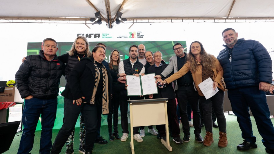 A fotografia em plano inteiro mostra um grupo numeroso de pessoas reunidas no centro de um palco sob uma estrutura de tenda branca, celebrando a assinatura de um ato oficial.

A&ccedil;&atilde;o Central: No centro do grupo, o governador Eduardo Leite segura uma pasta aberta de cor verde, exibindo um documento oficial com assinaturas. V&aacute;rias pessoas ao seu redor aproximam-se sorrindo para posar juntas para a foto. &Agrave; frente do governador, h&aacute; um pequeno balc&atilde;o ou p&uacute;lpito m&oacute;vel de cor clara.

Os Participantes:

O governador Eduardo Leite (ao centro) veste camisa branca sob um colete acolchoado preto com uma pequena bandeira do Rio Grande do Sul no peito, cal&ccedil;a escura e t&ecirc;nis claros.

&Agrave; sua direita (esquerda da foto), destacam-se uma mulher &agrave; frente usando um casaco preto com um cachecol estampado em tons de bege e preto, um homem na extremidade esquerda vestindo jaqueta acolchoada preta e cal&ccedil;a jeans, e uma mulher loira sorridente com colete acolchoado preto logo atr&aacute;s. Outras pessoas completam o grupo ao fundo.

&Agrave; sua esquerda (direita da foto), uma mulher com casaco marrom claro se inclina levemente segurando folhas de papel branco, ladeada por um homem de palet&oacute; cinza sobre camisa escura. Na extremidade direita, um homem robusto veste uma jaqueta acolchoada azul-marinho e cal&ccedil;a escura. Outros homens de trajes sociais e casuais acompanham o ato logo atr&aacute;s do p&uacute;lpito.

O Palco e Plano de Fundo:

O piso do palco &eacute; revestido por uma superf&iacute;cie verde que simula grama sint&eacute;tica.

Atr&aacute;s do grupo, h&aacute; um grande painel digital com fundo verde-brilhante.

Logo acima do painel, na lona branca da tenda, est&atilde;o aplicados os logotipos oficiais da CUFA (Central &Uacute;nica das Favelas), da Secretaria do Esporte e Lazer e do Governo do Estado do Rio Grande do Sul. No teto da estrutura, refletores de ilumina&ccedil;&atilde;o c&ecirc;nica aparecem fixados.

Na extrema esquerda, em segundo plano, nota-se uma bola de v&ocirc;lei ou futebol de cor amarela e azul sobre caixas de papel&atilde;o institucionais.

A imagem registra o momento de celebra&ccedil;&atilde;o e formaliza&ccedil;&atilde;o de parcerias entre o governo estadual, entidades sociais e representantes do esporte.