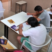 Foto mostra de cima uma sala onde h&aacute; apenados sentados em carteiras de sala de aula. Eles est&atilde;o lendo livros.