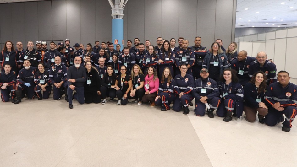 A fotografia em plano geral e &acirc;ngulo frontal mostra um grande grupo de profissionais de sa&uacute;de e socorristas reunidos de p&eacute; e agachados, posando juntos para uma foto coletiva em um amplo pavilh&atilde;o de eventos.

Os Integrantes: O grupo &eacute; composto por dezenas de homens e mulheres dispostos em duas fileiras principais (uma em p&eacute; ao fundo e outra agachada ou sentada &agrave; frente).

A grande maioria dos participantes veste o uniforme oficial do SAMU (Servi&ccedil;o de Atendimento M&oacute;vel de Urg&ecirc;ncia), que consiste em um macac&atilde;o ou conjunto de cor azul-marinho com faixas refletivas horizontais nas cores laranja e cinza nos bra&ccedil;os e pernas, al&eacute;m de patches com o logotipo do servi&ccedil;o e o n&uacute;mero 192. Alguns usam crach&aacute;s com cord&atilde;o verde pendurados no pesco&ccedil;o.

No centro da fileira da frente, destacam-se algumas pessoas com roupas civis e casuais: um homem de barba branca e blusa social escura, uma mulher com uma blusa rosa-choque brilhante e outras mulheres vestindo camisetas pretas ou blusas escuras.

O Ambiente:

O grupo est&aacute; posicionado no centro de um grande sal&atilde;o ou centro de conven&ccedil;&otilde;es com piso liso e claro em tom bege.

Ao fundo, h&aacute; paredes divis&oacute;rias modulares em tons de cinza-escuro e estruturas de estandes brancos &agrave; direita.

Uma pilastra cil&iacute;ndrica de cor azul-clara ergue-se pr&oacute;ximo ao centro, ramificando-se no topo em uma estrutura met&aacute;lica branca integrada ao teto do pavilh&atilde;o. A ilumina&ccedil;&atilde;o superior &eacute; ampla, uniforme e difusa.

A imagem registra o esp&iacute;rito de uni&atilde;o, integra&ccedil;&atilde;o e esp&iacute;rito de equipe entre os profissionais do servi&ccedil;o de atendimento de urg&ecirc;ncia do estado.