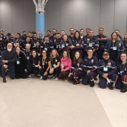 A fotografia em plano geral e &acirc;ngulo frontal mostra um grande grupo de profissionais de sa&uacute;de e socorristas reunidos de p&eacute; e agachados, posando juntos para uma foto coletiva em um amplo pavilh&atilde;o de eventos.

Os Integrantes: O grupo &eacute; composto por dezenas de homens e mulheres dispostos em duas fileiras principais (uma em p&eacute; ao fundo e outra agachada ou sentada &agrave; frente).

A grande maioria dos participantes veste o uniforme oficial do SAMU (Servi&ccedil;o de Atendimento M&oacute;vel de Urg&ecirc;ncia), que consiste em um macac&atilde;o ou conjunto de cor azul-marinho com faixas refletivas horizontais nas cores laranja e cinza nos bra&ccedil;os e pernas, al&eacute;m de patches com o logotipo do servi&ccedil;o e o n&uacute;mero 192. Alguns usam crach&aacute;s com cord&atilde;o verde pendurados no pesco&ccedil;o.

No centro da fileira da frente, destacam-se algumas pessoas com roupas civis e casuais: um homem de barba branca e blusa social escura, uma mulher com uma blusa rosa-choque brilhante e outras mulheres vestindo camisetas pretas ou blusas escuras.

O Ambiente:

O grupo est&aacute; posicionado no centro de um grande sal&atilde;o ou centro de conven&ccedil;&otilde;es com piso liso e claro em tom bege.

Ao fundo, h&aacute; paredes divis&oacute;rias modulares em tons de cinza-escuro e estruturas de estandes brancos &agrave; direita.

Uma pilastra cil&iacute;ndrica de cor azul-clara ergue-se pr&oacute;ximo ao centro, ramificando-se no topo em uma estrutura met&aacute;lica branca integrada ao teto do pavilh&atilde;o. A ilumina&ccedil;&atilde;o superior &eacute; ampla, uniforme e difusa.

A imagem registra o esp&iacute;rito de uni&atilde;o, integra&ccedil;&atilde;o e esp&iacute;rito de equipe entre os profissionais do servi&ccedil;o de atendimento de urg&ecirc;ncia do estado.