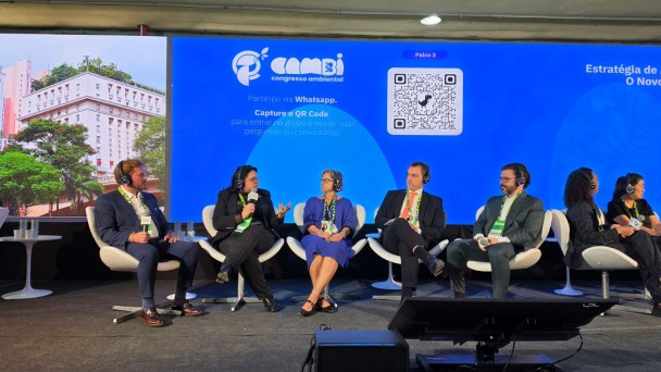 A imagem mostra um painel de discuss&atilde;o em um evento, com participantes sentados em palco usando fones e microfones, diante de um tel&atilde;o azul com informa&ccedil;&otilde;es do Congresso Ambiental.
