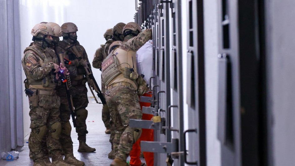 Corredor interno com paredes claras e ilumina&ccedil;&atilde;o homog&ecirc;nea, onde um grupo de agentes t&aacute;ticos usando uniformes camuflados, capacetes e equipamentos de prote&ccedil;&atilde;o conduz uma a&ccedil;&atilde;o em frente a uma fileira de portas met&aacute;licas de uma pris&atilde;o. 