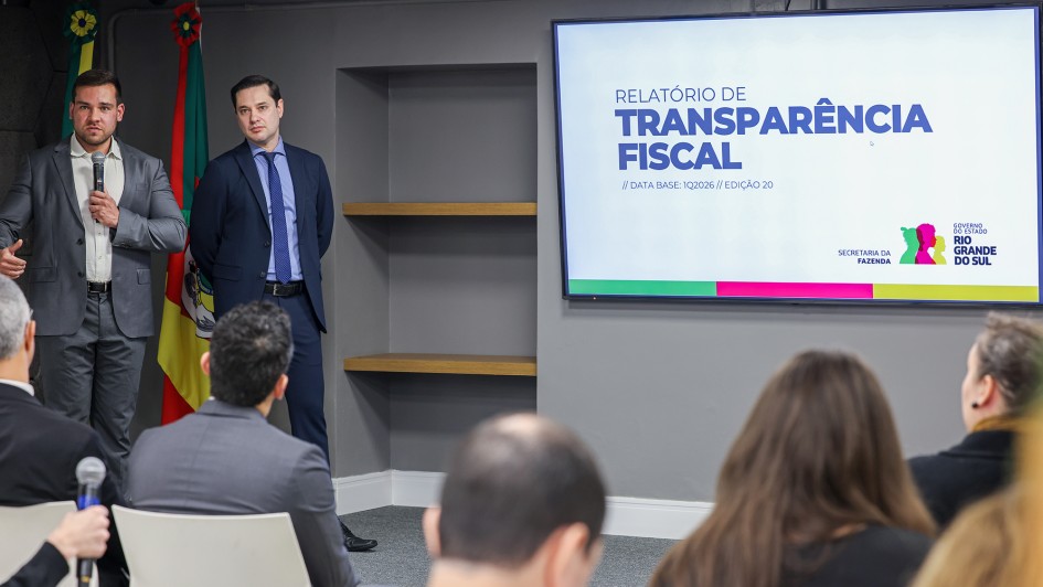 Sala de apresenta&ccedil;&atilde;o institucional com paredes neutras e ilumina&ccedil;&atilde;o uniforme, onde duas pessoas em trajes formais est&atilde;o em frente a um p&uacute;blico sentado. Ao lado, uma tela exibe o t&iacute;tulo &ldquo;RELAT&Oacute;RIO DE TRANSPAR&Ecirc;NCIA FISCAL // DATA BASE: 1Q2026 // EDI&Ccedil;&Atilde;O 20&rdquo;, acompanhado do logotipo do Governo do Estado do Rio Grande do Sul.