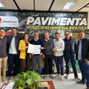 Ambiente interno de escrit&oacute;rio com ilumina&ccedil;&atilde;o de teto e mobili&aacute;rio simples, onde um grupo de sete pessoas posa lado a lado diante de um painel grande com a inscri&ccedil;&atilde;o &ldquo;PAVIMENTA &ndash; MUNICIPALISMO NA PR&Aacute;TICA&rdquo; e o logotipo do Governo do Estado do Rio Grande do Sul. No centro, duas pessoas seguram um documento aberto voltado para a c&acirc;mera.