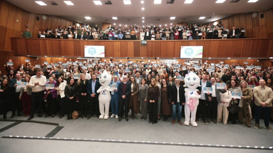 A fotografia em plano panor&acirc;mico e &acirc;ngulo frontal mostra uma grande celebra&ccedil;&atilde;o coletiva dentro de um amplo audit&oacute;rio lotado, com centenas de pessoas erguendo certificados azuis.

Primeiro Plano (Frente do Palco):

No centro da plateia, no n&iacute;vel do ch&atilde;o &agrave; frente das poltronas, um grupo de autoridades, prefeitos e representantes pol&iacute;ticos posa alinhado lado a lado. Eles vestem trajes sociais e casuais variados (ternos, blazers, camisas e vestidos).

Entre as autoridades, destacam-se dois mascotes em tamanho real baseados na figura do Z&eacute; Gotinha. O da esquerda &eacute; totalmente branco e traz o logotipo do SUS em azul no peito. O da direita representa uma vers&atilde;o feminina, usando um la&ccedil;o rosa na cabe&ccedil;a e segurando um beb&ecirc; de brinquedo envolto em uma manta rosa.

Segundo Plano (Plateia e Mezanino):

O audit&oacute;rio possui dois n&iacute;veis principais inteiramente ocupados pelo p&uacute;blico. O pavimento t&eacute;rreo e a galeria superior (mezanino ao fundo) est&atilde;o repletos de pessoas em p&eacute;.

Quase todas as pessoas da plateia seguram e exibem um certificado em formato retangular horizontal de cor azul-clara &agrave; altura do peito ou do rosto, criando um efeito visual de mosaico azul por todo o ambiente.

O Ambiente:

O audit&oacute;rio tem um design moderno com paredes revestidas por pain&eacute;is de madeira em tom castanho-claro.

Na parede divis&oacute;ria entre o t&eacute;rreo e o mezanino, est&atilde;o fixadas duas telas de TV digitais (uma &agrave; esquerda e outra &agrave; direita) que exibem um selo circular azul e rosa com o texto "MUNIC&Iacute;PIO AMIGO DA VACINA".

O teto possui diversas lumin&aacute;rias embutidas quadradas de luz branca, garantindo uma ilumina&ccedil;&atilde;o clara, homog&ecirc;nea e festiva para o registro fotogr&aacute;fico de grande escala.

A imagem registra o sucesso de uma pol&iacute;tica p&uacute;blica de sa&uacute;de e a mobiliza&ccedil;&atilde;o de munic&iacute;pios em torno da imuniza&ccedil;&atilde;o ga&uacute;cha.