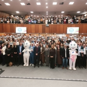 A fotografia em plano panor&acirc;mico e &acirc;ngulo frontal mostra uma grande celebra&ccedil;&atilde;o coletiva dentro de um amplo audit&oacute;rio lotado, com centenas de pessoas erguendo certificados azuis.

Primeiro Plano (Frente do Palco):

No centro da plateia, no n&iacute;vel do ch&atilde;o &agrave; frente das poltronas, um grupo de autoridades, prefeitos e representantes pol&iacute;ticos posa alinhado lado a lado. Eles vestem trajes sociais e casuais variados (ternos, blazers, camisas e vestidos).

Entre as autoridades, destacam-se dois mascotes em tamanho real baseados na figura do Z&eacute; Gotinha. O da esquerda &eacute; totalmente branco e traz o logotipo do SUS em azul no peito. O da direita representa uma vers&atilde;o feminina, usando um la&ccedil;o rosa na cabe&ccedil;a e segurando um beb&ecirc; de brinquedo envolto em uma manta rosa.

Segundo Plano (Plateia e Mezanino):

O audit&oacute;rio possui dois n&iacute;veis principais inteiramente ocupados pelo p&uacute;blico. O pavimento t&eacute;rreo e a galeria superior (mezanino ao fundo) est&atilde;o repletos de pessoas em p&eacute;.

Quase todas as pessoas da plateia seguram e exibem um certificado em formato retangular horizontal de cor azul-clara &agrave; altura do peito ou do rosto, criando um efeito visual de mosaico azul por todo o ambiente.

O Ambiente:

O audit&oacute;rio tem um design moderno com paredes revestidas por pain&eacute;is de madeira em tom castanho-claro.

Na parede divis&oacute;ria entre o t&eacute;rreo e o mezanino, est&atilde;o fixadas duas telas de TV digitais (uma &agrave; esquerda e outra &agrave; direita) que exibem um selo circular azul e rosa com o texto "MUNIC&Iacute;PIO AMIGO DA VACINA".

O teto possui diversas lumin&aacute;rias embutidas quadradas de luz branca, garantindo uma ilumina&ccedil;&atilde;o clara, homog&ecirc;nea e festiva para o registro fotogr&aacute;fico de grande escala.

A imagem registra o sucesso de uma pol&iacute;tica p&uacute;blica de sa&uacute;de e a mobiliza&ccedil;&atilde;o de munic&iacute;pios em torno da imuniza&ccedil;&atilde;o ga&uacute;cha.