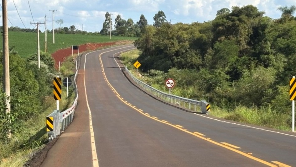 Esta &eacute; uma descri&ccedil;&atilde;o da imagem "Cerro Largo e Rolador 1.jpg" para fins de acessibilidade:

A fotografia em plano geral e perspectiva linear mostra uma rodovia asfaltada e sinuosa que corta uma paisagem rural em um dia claro.

A Rodovia: A estrada de asfalto escuro e novo ocupa grande parte da metade inferior e central do quadro. Ela se estende em dire&ccedil;&atilde;o ao fundo, fazendo uma curva suave para a direita e, em seguida, uma inflex&atilde;o para a esquerda, acompanhando o relevo ondulado. A pista &eacute; dividida por uma linha amarela tracejada dupla no centro e delimitada por linhas brancas cont&iacute;nuas nas bordas.

Sinaliza&ccedil;&atilde;o e Seguran&ccedil;a:

Nas laterais da pista, em trechos onde h&aacute; declives nas margens, est&atilde;o instaladas barreiras met&aacute;licas de prote&ccedil;&atilde;o (guardrails) de cor prata. Nas extremidades dessas barreiras, h&aacute; placas verticais listradas em diagonal com as cores amarelo e preto.

&Agrave; direita da estrada, erguem-se duas placas de tr&acirc;nsito verticais: uma placa amarela em formato de losango indicando uma curva &agrave; frente e, logo depois, uma placa circular branca com borda vermelha e uma tarja diagonal, indicando proibi&ccedil;&atilde;o de ultrapassagem.

A Paisagem e o Entorno:

Do lado esquerdo da rodovia, h&aacute; um terreno plano coberto por uma planta&ccedil;&atilde;o verde e homog&ecirc;nea (campo agr&iacute;cola), margeado por uma fileira de postes de energia el&eacute;trica de concreto. Pr&oacute;ximo ao in&iacute;cio da curva, nota-se uma pequena encosta de terra de cor vermelha exposta.

Do lado direito, estende-se uma &aacute;rea de vegeta&ccedil;&atilde;o nativa mais densa, composta por &aacute;rvores e arbustos de copas verdes e variadas.

O C&eacute;u: Na linha do horizonte, o c&eacute;u aparece claro com algumas nuvens brancas dispersas e volumosas, indicando condi&ccedil;&otilde;es de tempo bom.

A imagem registra a infraestrutura de transporte rodovi&aacute;rio e as condi&ccedil;&otilde;es de sinaliza&ccedil;&atilde;o em uma &aacute;rea de interior.