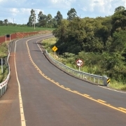 Esta &eacute; uma descri&ccedil;&atilde;o da imagem "Cerro Largo e Rolador 1.jpg" para fins de acessibilidade:

A fotografia em plano geral e perspectiva linear mostra uma rodovia asfaltada e sinuosa que corta uma paisagem rural em um dia claro.

A Rodovia: A estrada de asfalto escuro e novo ocupa grande parte da metade inferior e central do quadro. Ela se estende em dire&ccedil;&atilde;o ao fundo, fazendo uma curva suave para a direita e, em seguida, uma inflex&atilde;o para a esquerda, acompanhando o relevo ondulado. A pista &eacute; dividida por uma linha amarela tracejada dupla no centro e delimitada por linhas brancas cont&iacute;nuas nas bordas.

Sinaliza&ccedil;&atilde;o e Seguran&ccedil;a:

Nas laterais da pista, em trechos onde h&aacute; declives nas margens, est&atilde;o instaladas barreiras met&aacute;licas de prote&ccedil;&atilde;o (guardrails) de cor prata. Nas extremidades dessas barreiras, h&aacute; placas verticais listradas em diagonal com as cores amarelo e preto.

&Agrave; direita da estrada, erguem-se duas placas de tr&acirc;nsito verticais: uma placa amarela em formato de losango indicando uma curva &agrave; frente e, logo depois, uma placa circular branca com borda vermelha e uma tarja diagonal, indicando proibi&ccedil;&atilde;o de ultrapassagem.

A Paisagem e o Entorno:

Do lado esquerdo da rodovia, h&aacute; um terreno plano coberto por uma planta&ccedil;&atilde;o verde e homog&ecirc;nea (campo agr&iacute;cola), margeado por uma fileira de postes de energia el&eacute;trica de concreto. Pr&oacute;ximo ao in&iacute;cio da curva, nota-se uma pequena encosta de terra de cor vermelha exposta.

Do lado direito, estende-se uma &aacute;rea de vegeta&ccedil;&atilde;o nativa mais densa, composta por &aacute;rvores e arbustos de copas verdes e variadas.

O C&eacute;u: Na linha do horizonte, o c&eacute;u aparece claro com algumas nuvens brancas dispersas e volumosas, indicando condi&ccedil;&otilde;es de tempo bom.

A imagem registra a infraestrutura de transporte rodovi&aacute;rio e as condi&ccedil;&otilde;es de sinaliza&ccedil;&atilde;o em uma &aacute;rea de interior.