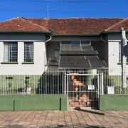 Fachada frontal de um pr&eacute;dio baixo com estilo simples e sim&eacute;trico, paredes claras com detalhes em verde e um telhado inclinado de telhas cer&acirc;micas avermelhadas; o acesso central possui uma escadaria coberta e &eacute; protegido por uma grade met&aacute;lica branca.