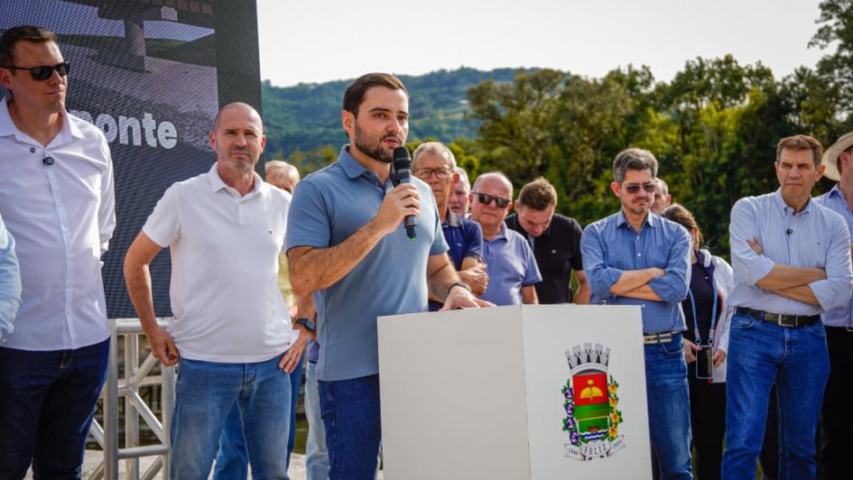 V&aacute;rias pessoasl est&atilde;o em p&eacute; em um ambiente externo, uma delas fala com microfone apoiada em um p&uacute;lpito branco com o bras&atilde;o em uma das fases
