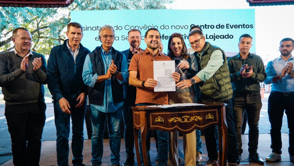 V&aacute;rias pessoas est&aacute; em p&eacute; atr&aacute;s de uma pequena mesa, segurando um documento assinado