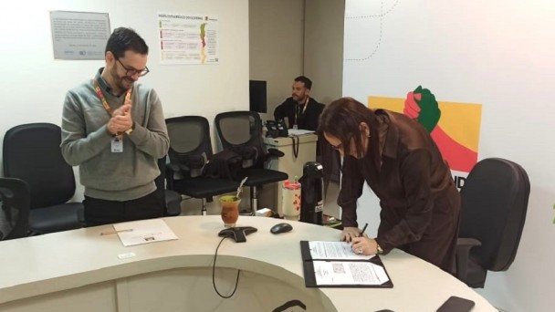 Ambiente de escrit&oacute;rio com balc&atilde;o identificado por &ldquo;RS&rdquo;, onde uma pessoa assina um documento enquanto outra observa ao lado.