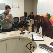 Ambiente de escrit&oacute;rio com balc&atilde;o identificado por &ldquo;RS&rdquo;, onde uma pessoa assina um documento enquanto outra observa ao lado.