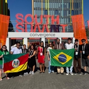 Grupo de pessoas posando em &aacute;rea externa diante de uma estrutura com o letreiro &ldquo;SOUTH SUMMIT&rdquo; e &ldquo;convergence&rdquo;, segurando bandeiras do Brasil e do Rio Grande do Sul; o local tem estruturas modernas com pain&eacute;is coloridos e circula&ccedil;&atilde;o de outras pessoas ao fundo.