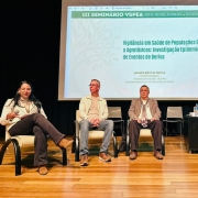 Foto mostra cinco debatedores, sentados em poltronas no palco do evento. No alto, uma tela projeta o nome do evento.