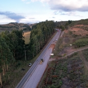 Vista a&eacute;rea de uma rodovia em &aacute;rea rural com vegeta&ccedil;&atilde;o e morros ao redor, onde um trecho est&aacute; em obras com m&aacute;quinas e ve&iacute;culos, incluindo caminh&otilde;es e equipamentos ao longo da pista parcialmente preparada.